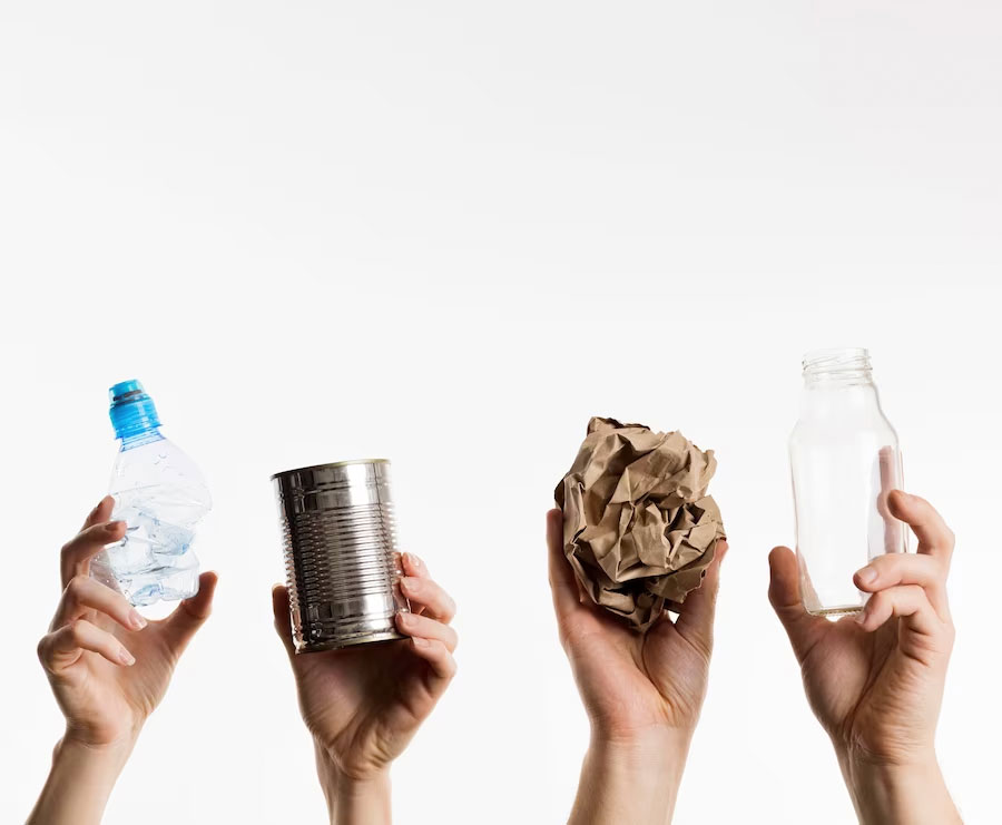 four hands holding up different objects to represent recyclable materials, a plastic water-bottle, a tin can, paper and a glass-bottle