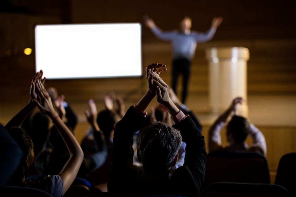 back view of an audience clapping in a professional conference with a speaker standing on a stage in the background with his arms up in the air.  