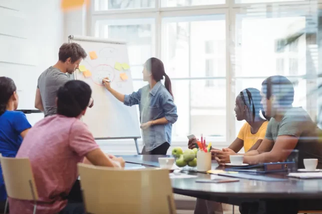 una sala de oficina con varias personas sentadas alrededor de una mesa mirando una pizarra blanca, donde una mujer está escribiendo una estrategia para comercializar cursos de capacitación.