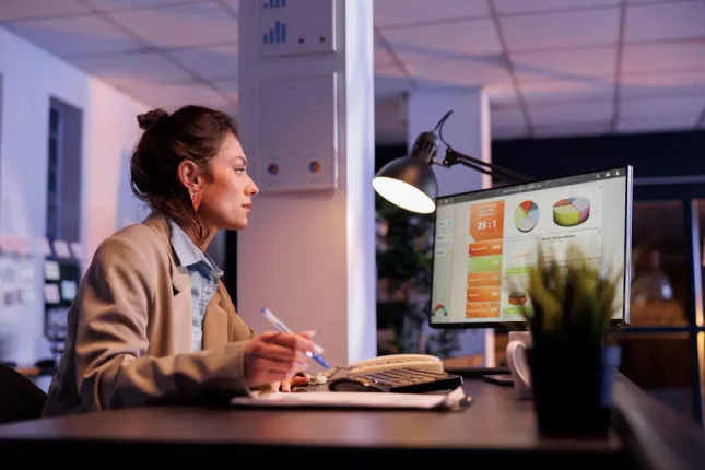 Una mujer sentada en un escritorio de trabajo frente a la pantalla de una computadora, mirando gráficos, muy concentrada y tomando notas.