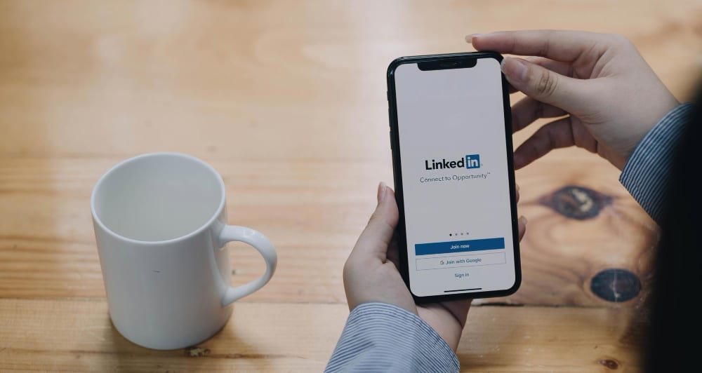 Close-up shot of two female hands holding a smartphone with the screen showing the login page for linkedin, with a white empty mug on the side.