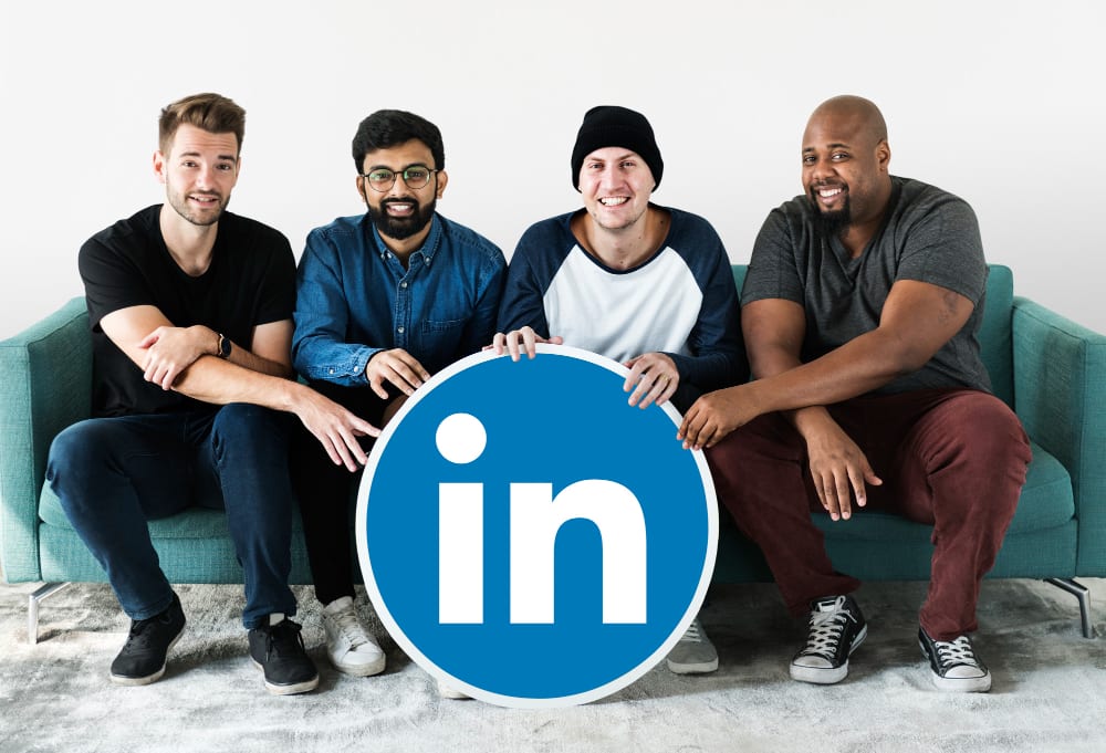 Four men seating on a couch holding a sign with the LinkedIn logo on it.