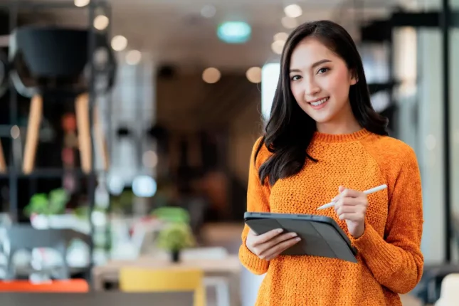 Una mujer asiática con un suéter naranja brillante, sonriendo y tomando notas en una tableta organizando su estrategia para comercializar programas de capacitación.