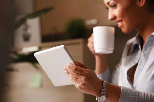 Een vrouw met een donkere huidskleur die een kopje koffie in de ene hand en een tablet in de andere hand houdt terwijl ze naar inhoud kijkt