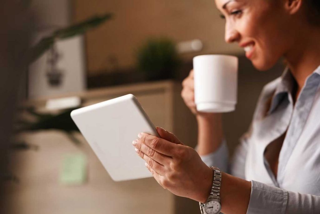 A dark-skinned woman holding a cup of coffee in one hand and a tablet in the other, smiling while watching content