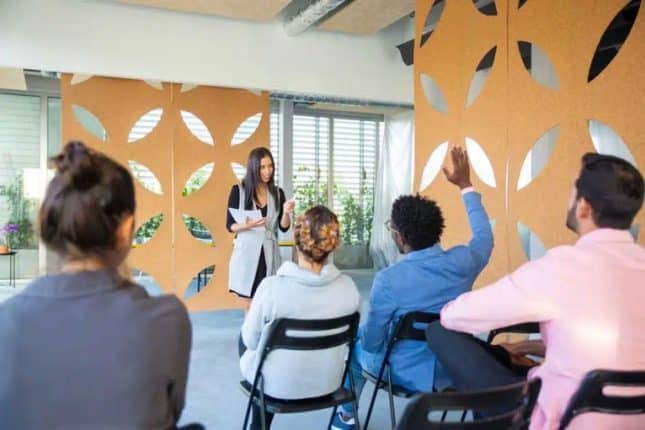Een vrouw leidt een persoonlijke trainingssessie in een prachtige ruimte en een persoon in het publiek steekt zijn hand op om een vraag te stellen
