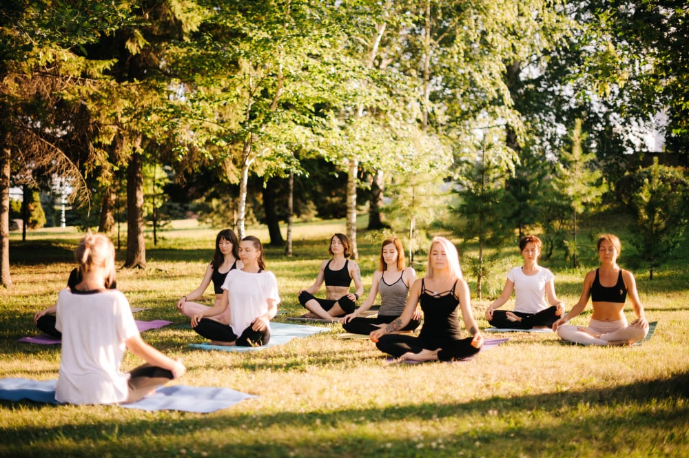 a yoga class event happening at a park on a sunny day with several women in a seated meditative position.