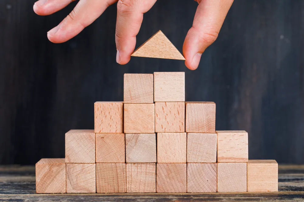 a hand placing the top portion of a pyramid formed by wooden blocks. Representing the conclusion of the cone of learning