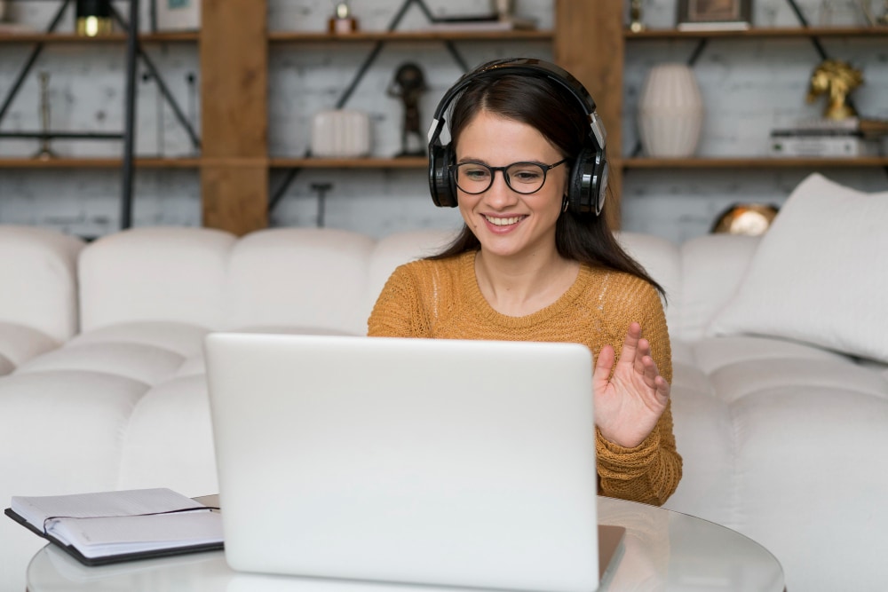 A young white woman with dark hair, wearing headphones and smiling while working on her laptop, with her notebook open by her side.