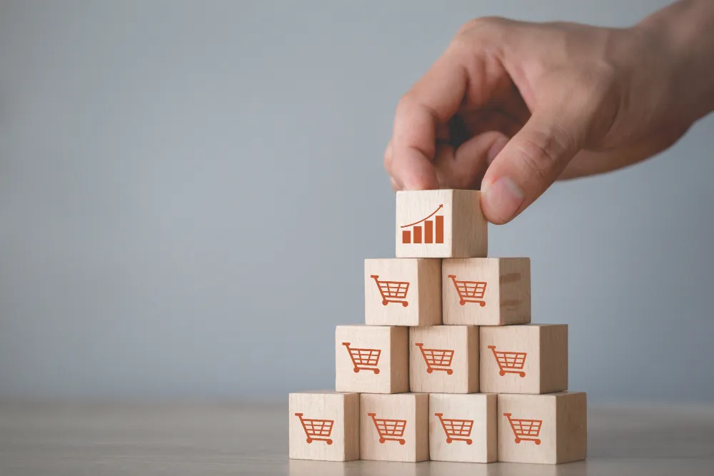 a male hand placing a small wooden block with the picture of a growth graph on it, on top of a pile of wooden blocks with images of shopping carts on it to represent how to sell courses online successfully.