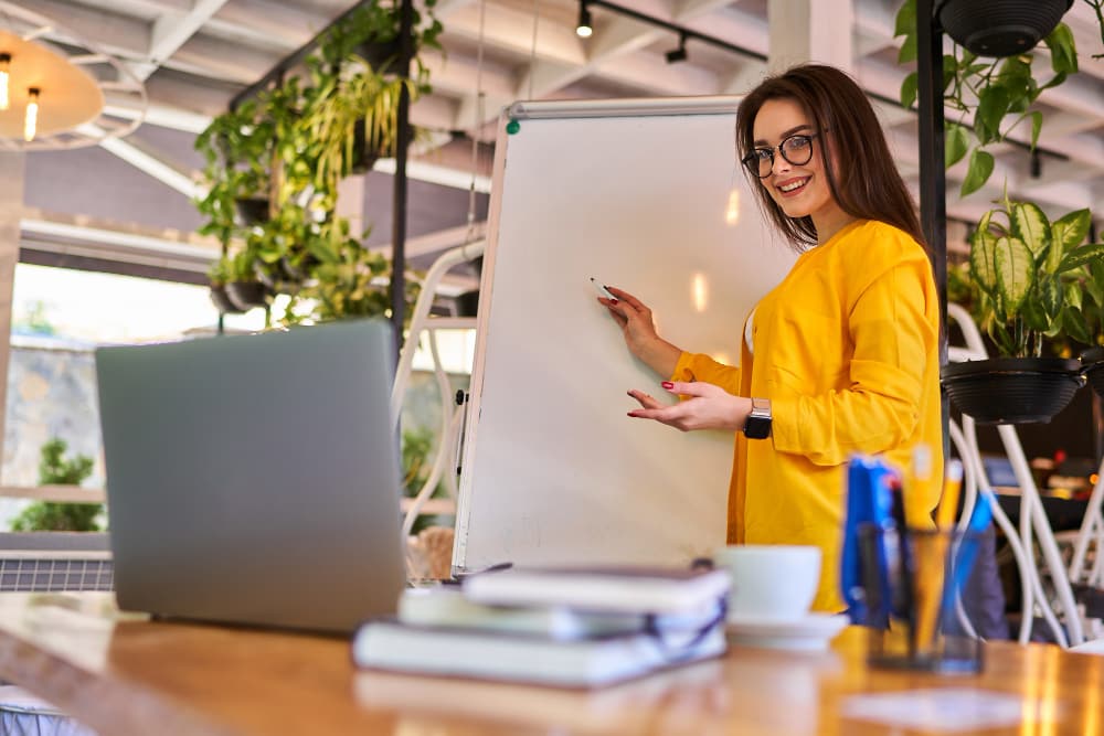 professional woman smiling wearing a yellow overall, standing in front of a whiteboard and lecturing towards a laptop.