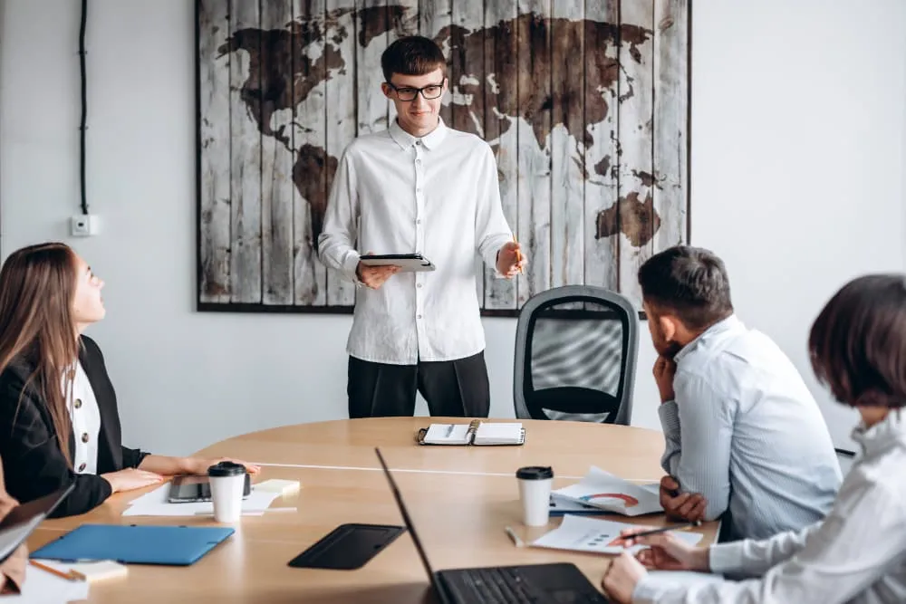 A young professional man standing at the edge of an office table while colleagues are sitting down and listening to him deliver a hybrid virtual instructor led training session.