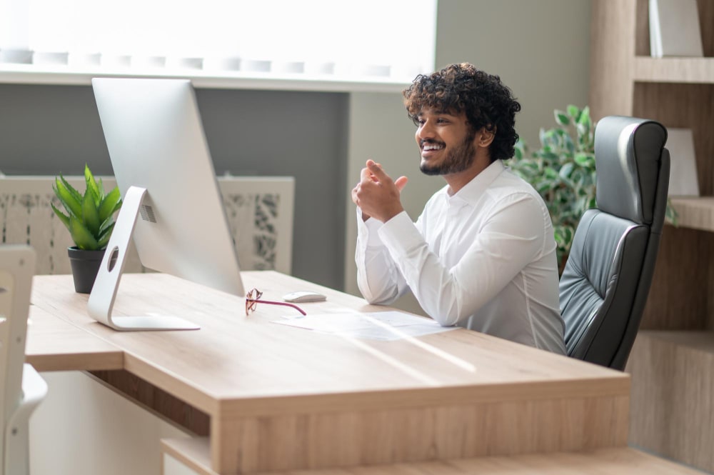 a young male professional sitting at a desk smiling in front of a computer screen.