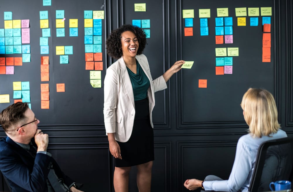 Een zwarte, middelbare vrouw staat op kantoor, lacht en is creatief bezig terwijl ze met haar team een ​​brainstormsessie houdt over de planning van evenementenmarketing.