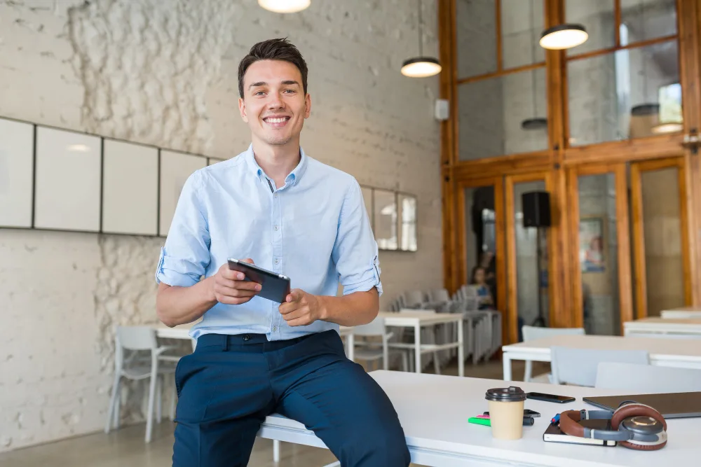 Joven profesional en un entorno de oficina, apoyado en una mesa mientras sostiene una tableta con el calendario de eventos de la comunidad en línea.